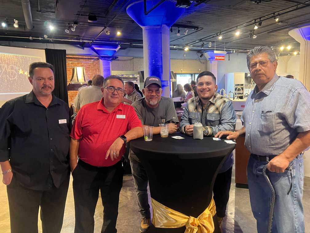 Five men standing around a black cocktail table with gold sash.