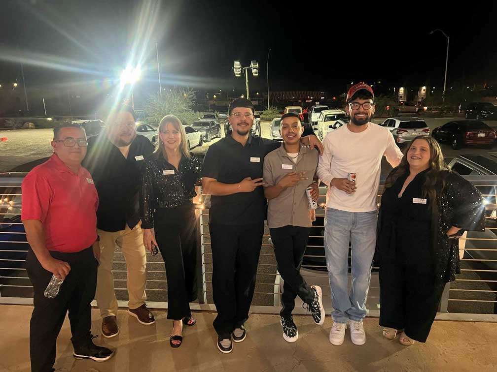 Group of six people smiling and posing outdoors in the parking lot at night.