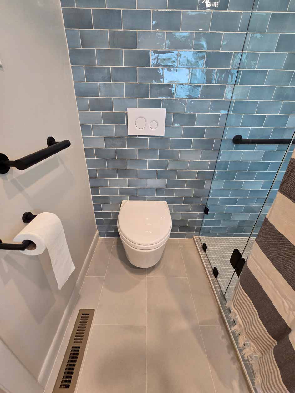 Minimalist bathroom with wall-mounted toilet, black grab bars, and blue subway tile wall next to glass shower