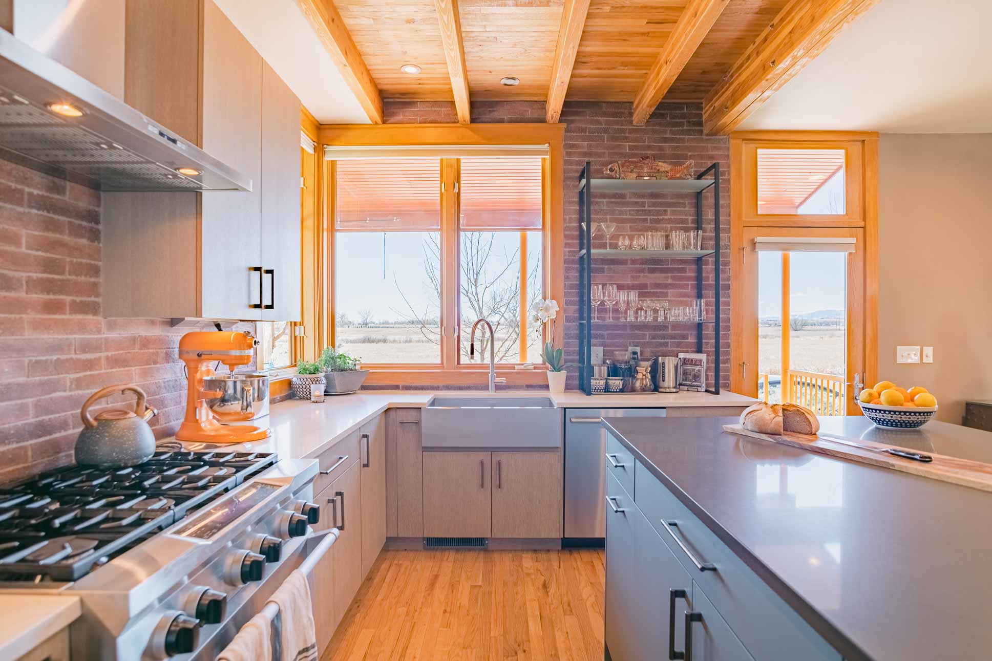 Close-up of kitchen sink under corner windows, with modern cabinets, brick walls, and open shelving
