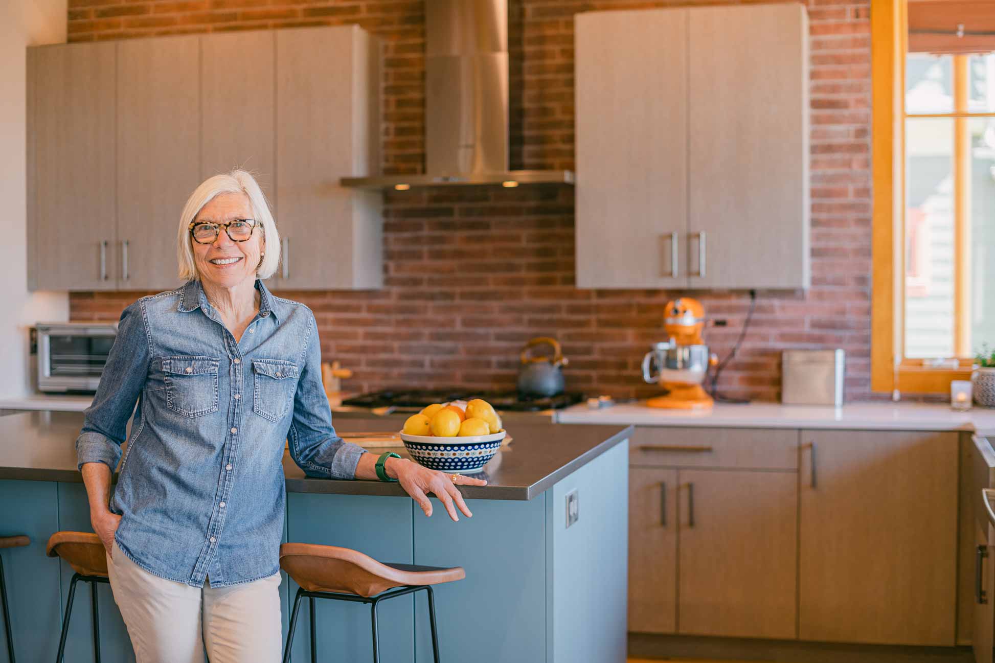 Smiling woman in denim shirt standing in a bright kitchen with exposed brick and light wood cabinetry