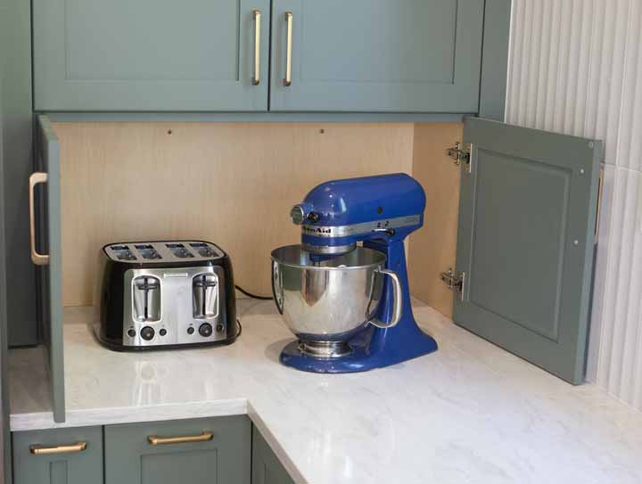 Appliance garage with sage green cabinet doors revealing a chrome toaster and blue stand mixer on a marble counter