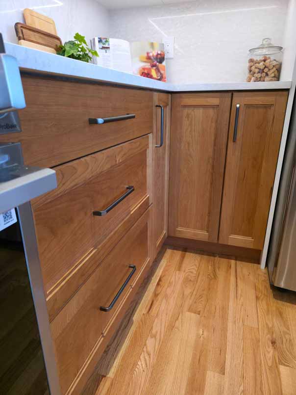 Close-up view of wooden kitchen cabinetry with dark handles, white countertops, hardwood flooring, and small potted herb plant on the counter.