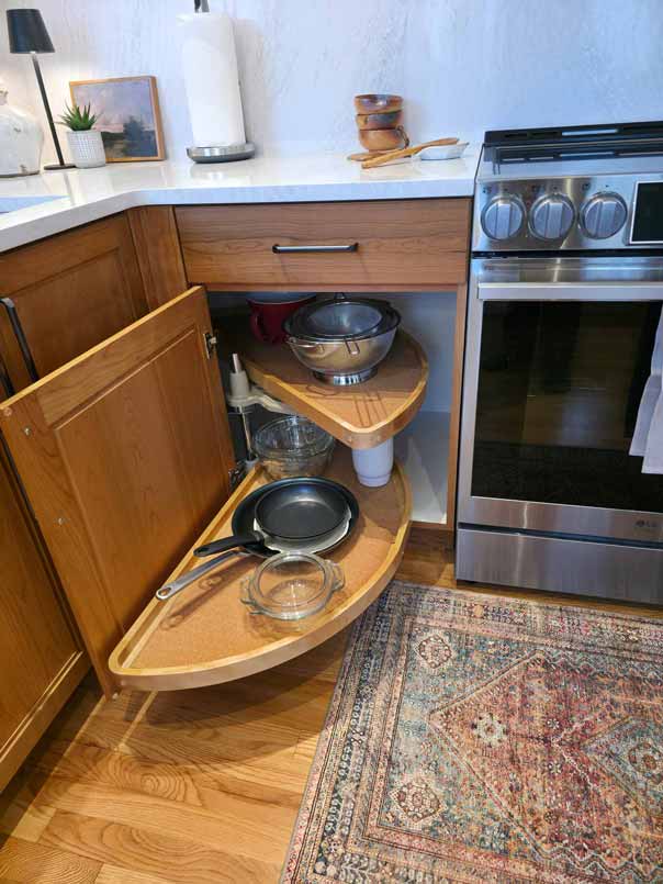 Kitchen corner cabinet with pull-out curved wooden shelves storing cookware including metal bowls, pans, and glass containers, next to a stainless steel stove and featuring white countertops with hardwood flooring and a decorative area rug.