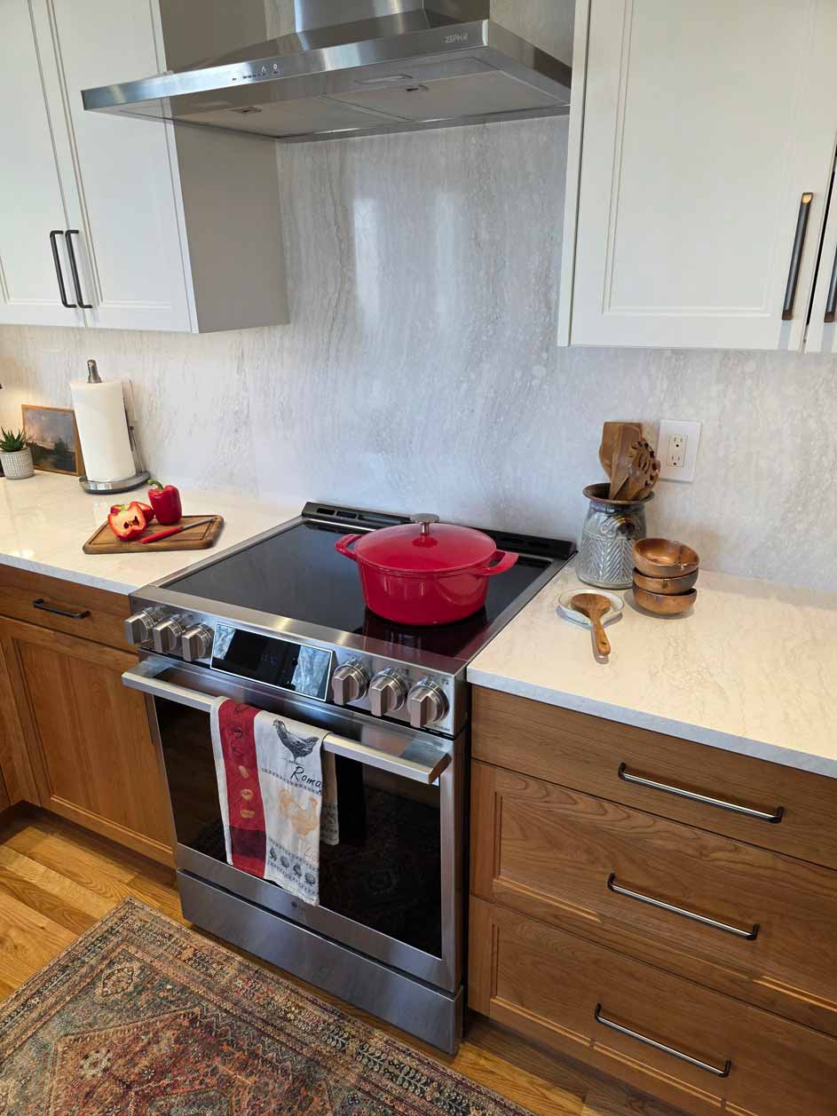 Close-up of kitchen cooktop with bright red Dutch oven, stainless steel range hood, white upper cabinets, wooden lower cabinets, white quartz countertops, marble-look backsplash, and cooking utensils displayed in a ceramic holder.