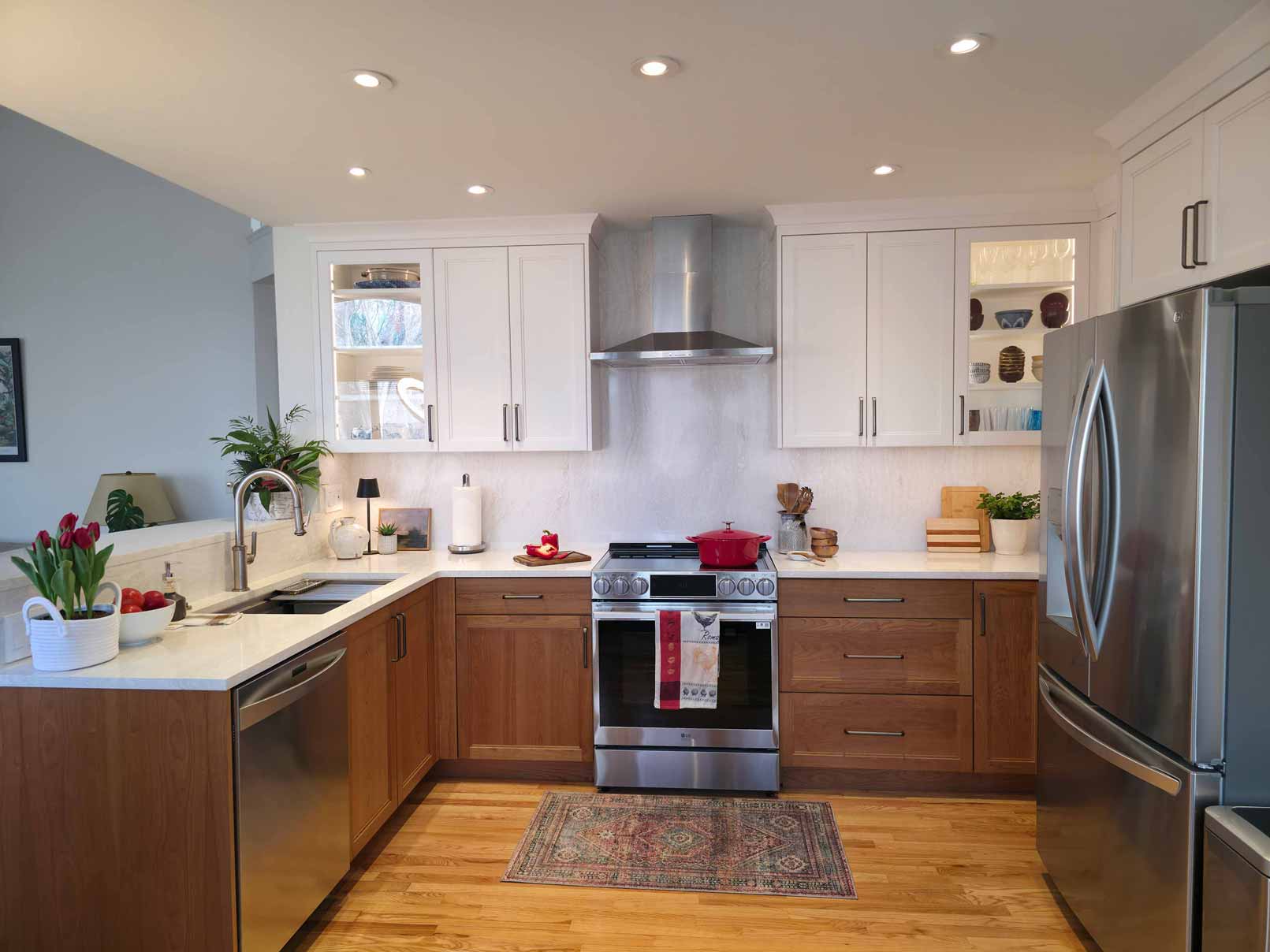 Full view of modern L-shaped kitchen with two-tone cabinetry featuring white upper cabinets and wooden lower cabinets, white countertops, stainless steel appliances, recessed lighting, display shelving, red cooking pot on stove, potted tulips by the sink, and hardwood flooring with an area rug.