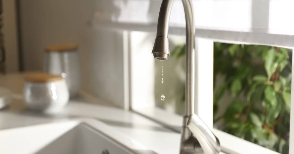 A close-up of a dripping kitchen faucet with water drops actively falling. The window next to it is open.