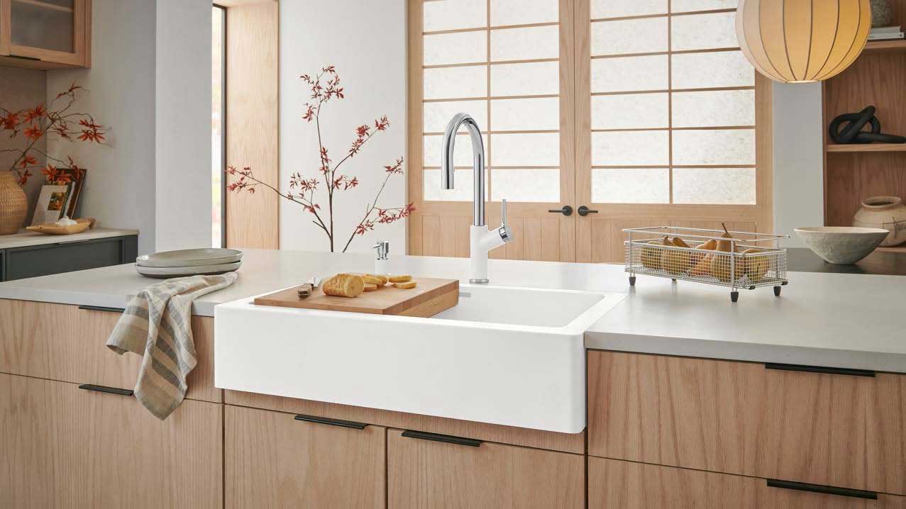A white apron-front sink is set into light wood cabinets in a modern kitchen with a Japandi aesthetic. A tall chrome and white faucet arches over the sink, and the background features a shoji-style door and a delicate branch wall decal.