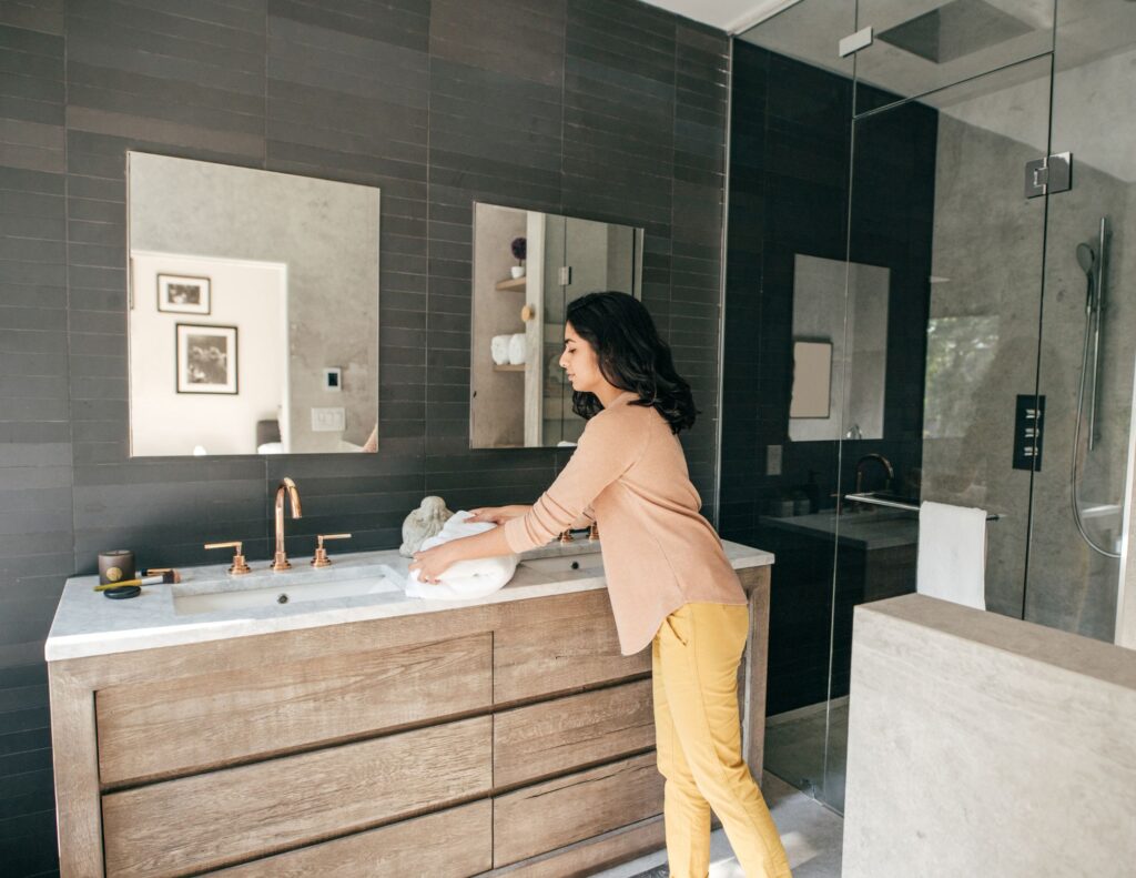 Woman cleaning a modern bathroom vanity with double sinks, wood cabinetry, and matte black tile wall.