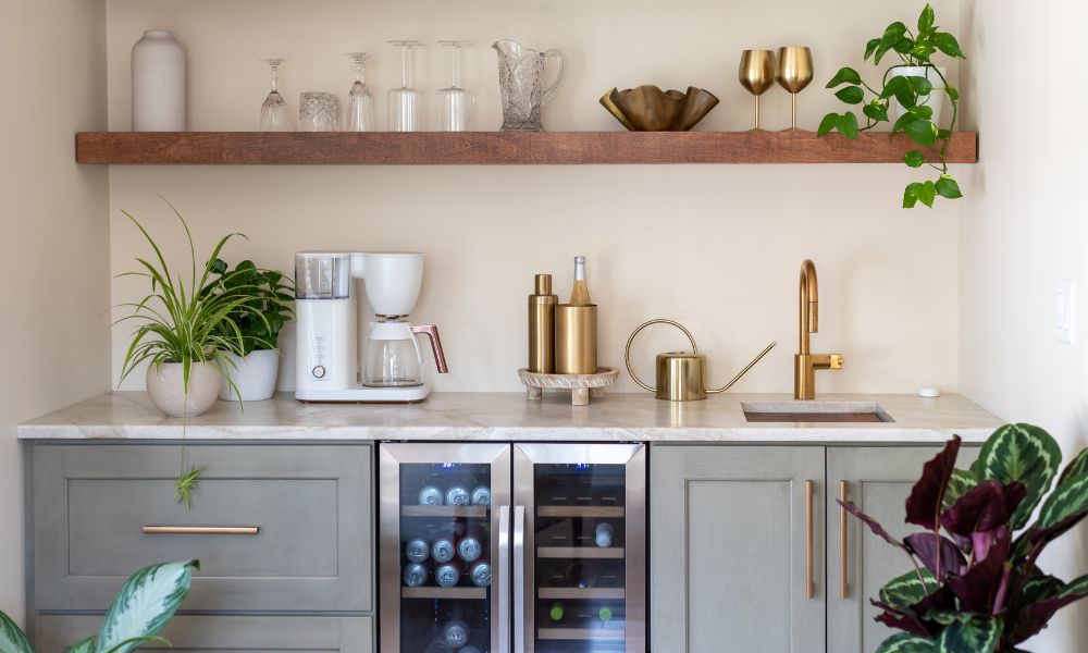 A contemporary coffee and wine bar with sage green cabinets, a built-in beverage fridge, a white countertop, and a floating wood shelf holding glasses and plants.