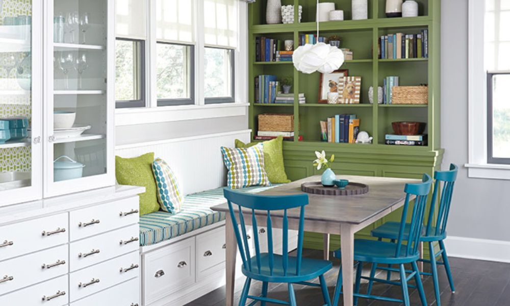 A bright breakfast nook featuring a large green built-in bookshelf, a white storage bench with colorful cushions, and vibrant blue chairs around a gray wooden table.