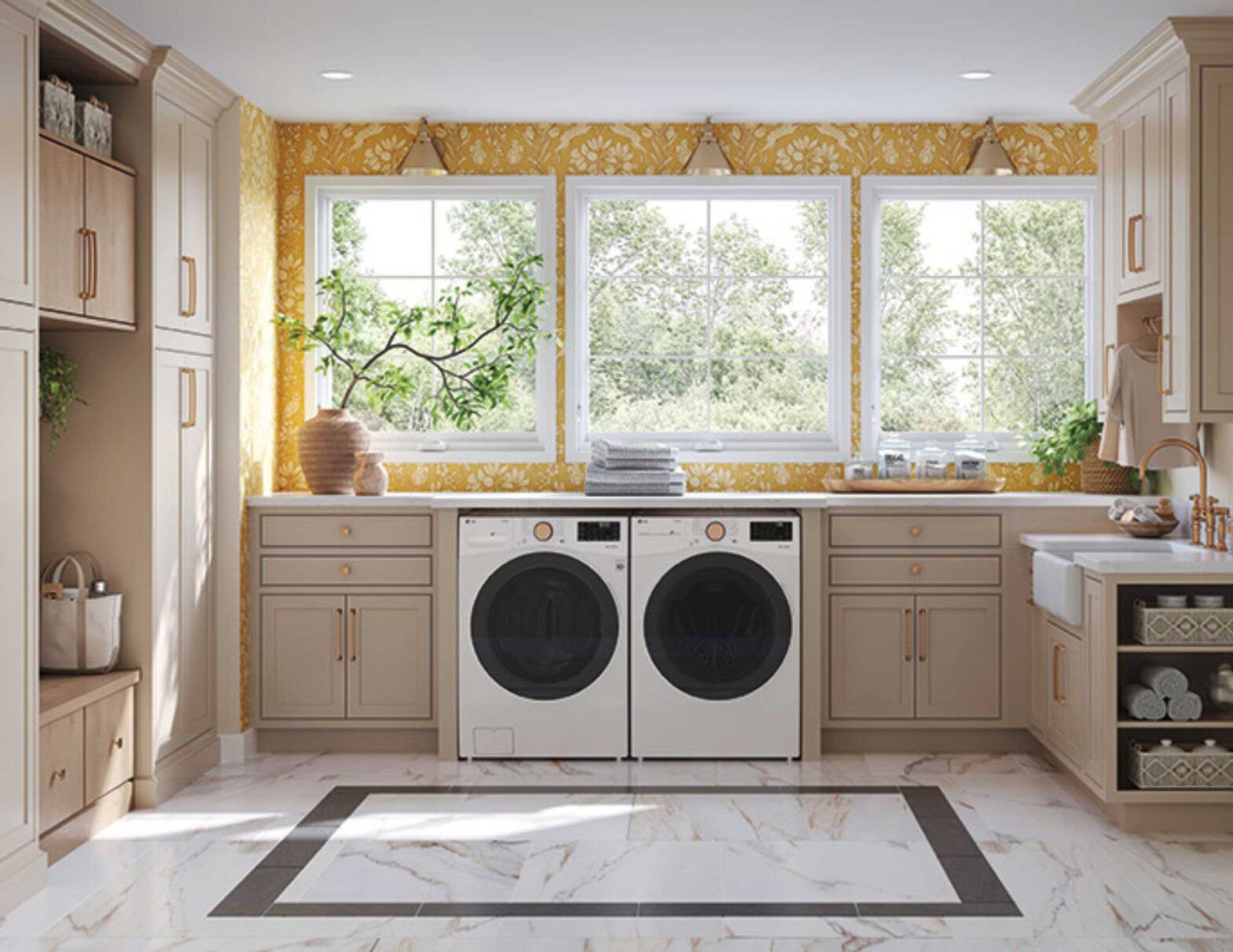 A sunny laundry room with light taupe cabinets, a bold yellow patterned wallpaper, and a front-loading washer and dryer set placed under a countertop between three large windows.