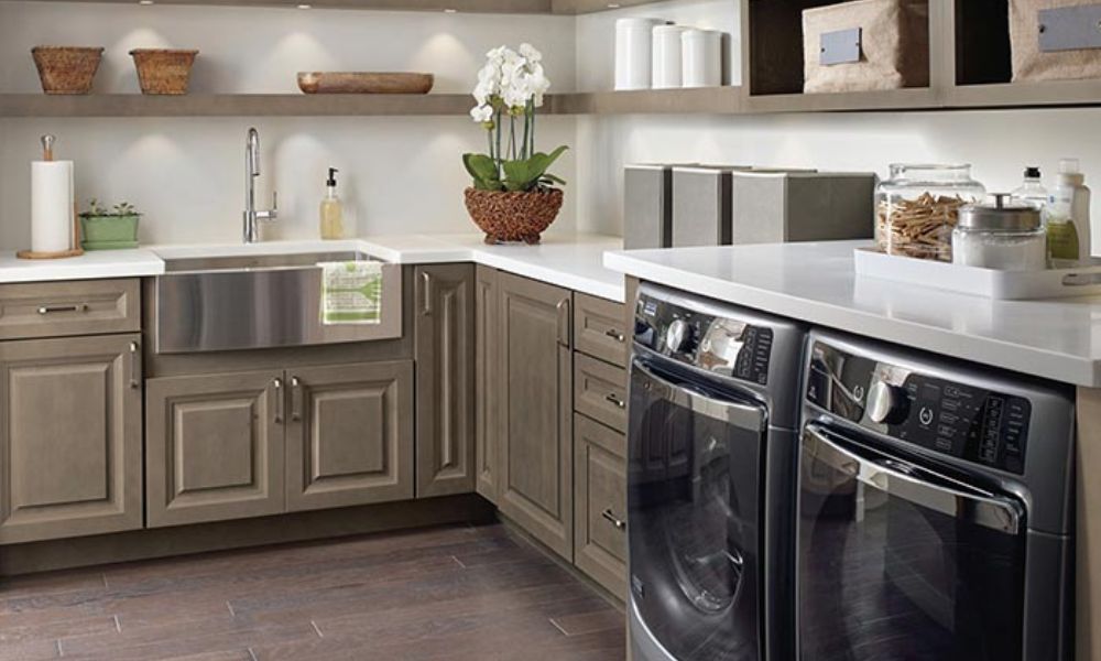 A sophisticated laundry room featuring taupe-brown raised-panel cabinets, a large stainless steel farmhouse sink, and a modern front-loading washer and dryer.