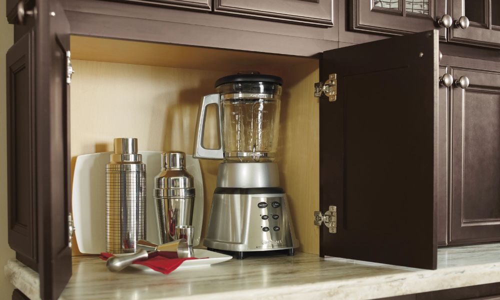 A dark wood appliance garage cabinet sitting on a countertop with doors open to reveal a blender and shaker set