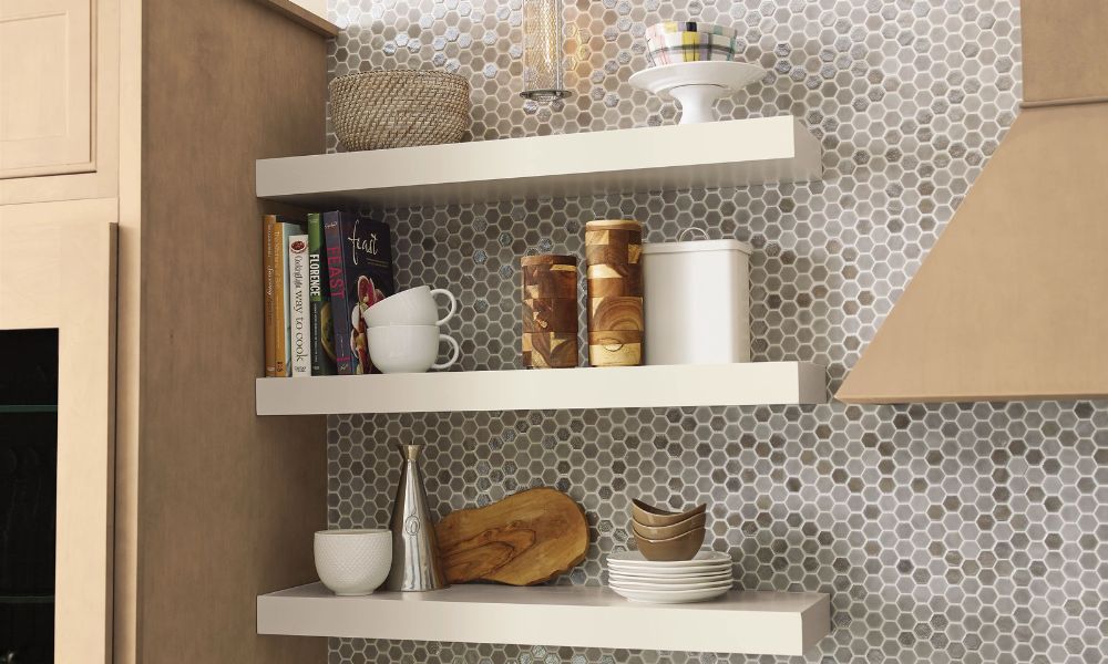 Three white floating shelves mounted on a tiled wall displaying bowls, jars, and kitchen decor