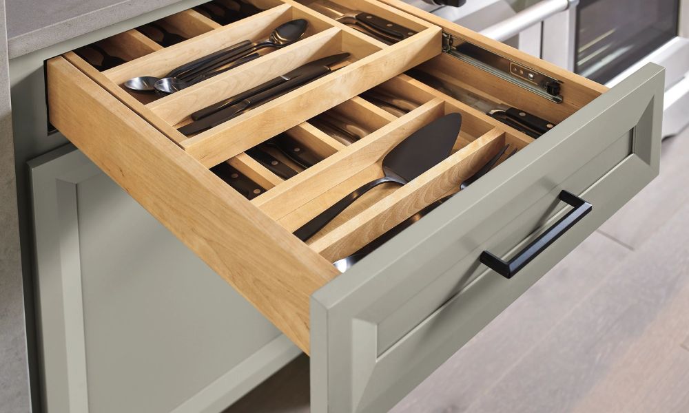 A drawer open to reveal a two-tiered wooden cutlery divider filled with silverware and serving utensils