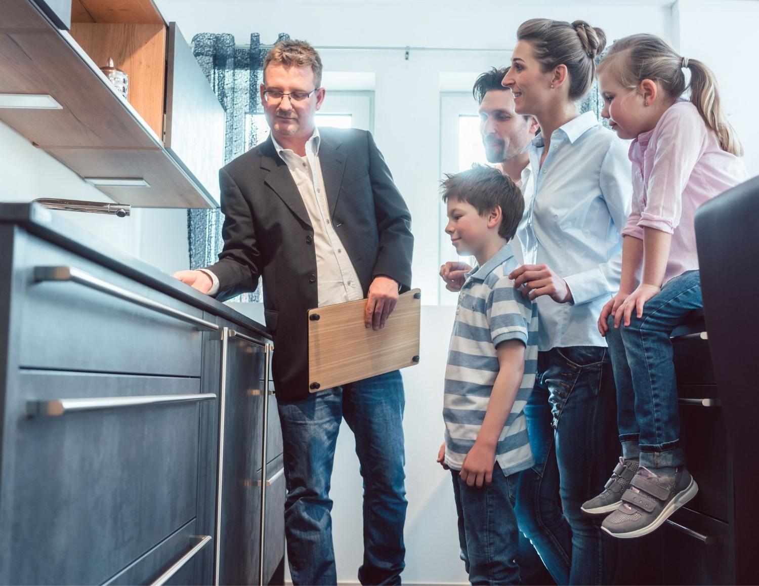 A family with two children is in a kitchen, attentively observing a man in a blazer demonstrating a cabinet feature. The atmosphere is informative and engaged.