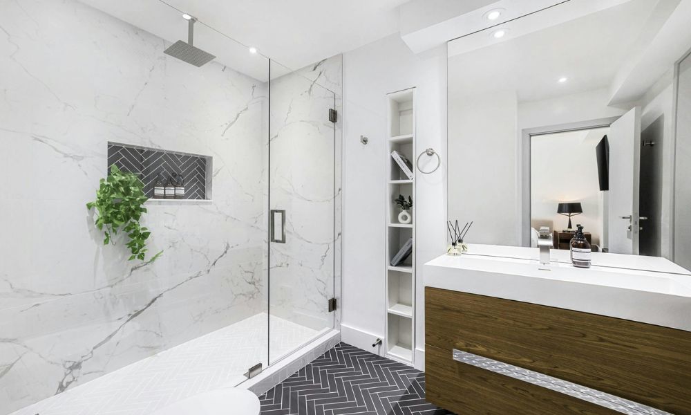 A bright modern bathroom with a marble-tiled walk-in shower, dark herringbone tile flooring, and a floating dark wood vanity with white quartz countertop
