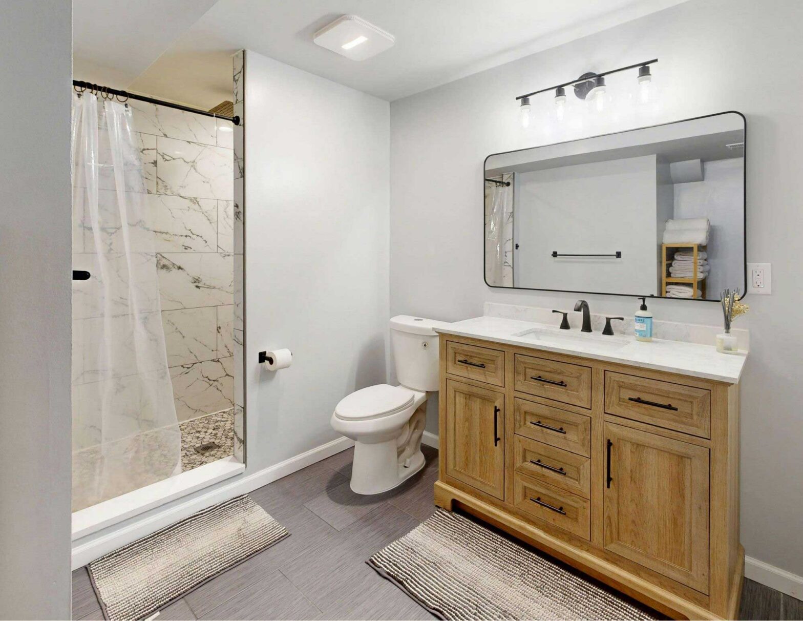 A finished basement bathroom featuring a walk-in marble-tiled shower with curtain, a natural wood vanity with white countertop, large rounded mirror, and matte black fixtures