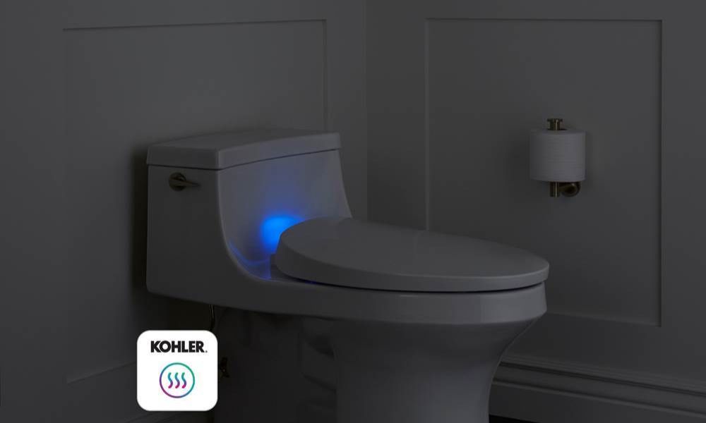 A one-piece toilet with an illuminated Kohler heated seat glowing blue in a dimly lit dark gray bathroom