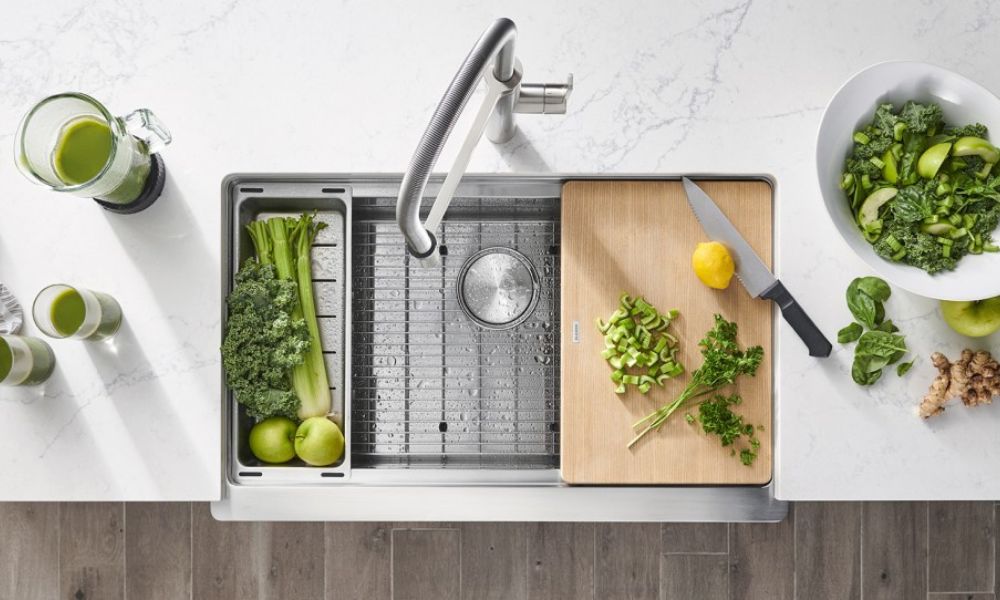 A kitchen sink with a cutting board and assorted fresh vegetables arranged beside it.