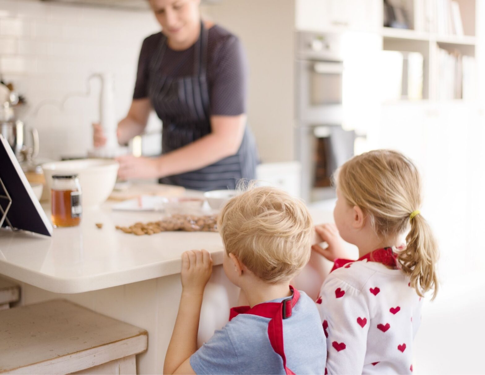 Two young children in red aprons peek over a white kitchen countertop while a parent prepares food